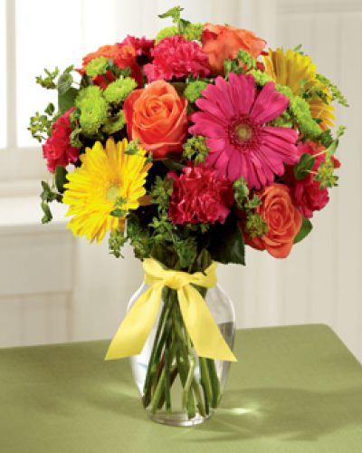 Celebrating life with colorful blooms that inspire and delight, this flower bouquet is ready to create a happy moment for your recipient that they will never forget. Orange roses, hot pink gerbera daisies, yellow gerbera daisies, hot pink carnations, green button poms, bupleurum, and lush greens mingle together to create a sunlit display while seated in a classic clear glass vase tied at the neck with a yellow satin ribbon for a sweet effect.
