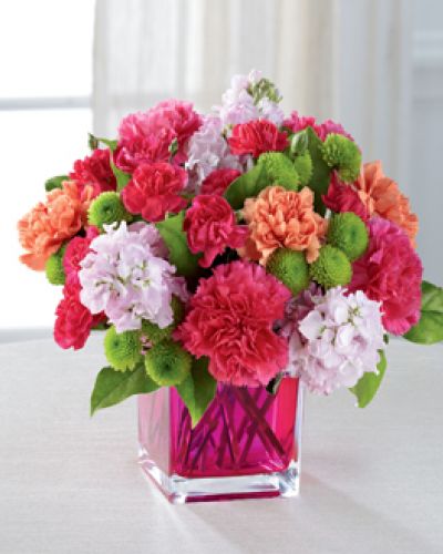 Dazzling and delightful, this bold and colorful flower bouquet is set to impress with it's high impact look and graceful styling. Hot pink carnations, orange carnations, pale pink gilly flower, hot pink mini carnations, green button poms, and lush greens are beautifully arranged in a raspberry pink glass cubed vase to create a rush of fun and warm wishes to send straight to your recipient's door.